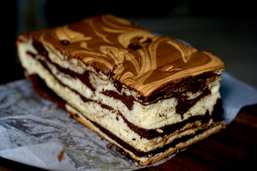 A rectangular cake with layered textures, featuring a marbled design of chocolate and cream-colored sections. It rests on parchment paper on a dark surface, and the cake's top shows a swirling pattern.