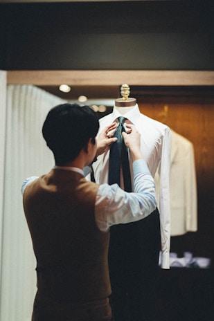 A person wearing a brown vest adjusts a dark tie on a mannequin dressed in a white shirt, situated in a well-lit tailor shop with wooden and cloth decor.