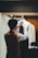 A person wearing a brown vest adjusts a dark tie on a mannequin dressed in a white shirt, situated in a well-lit tailor shop with wooden and cloth decor.