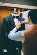 A tailor is adjusting a pinstripe suit on a mannequin. The suit is black with white pinstripes and is complemented by a black bow tie. The tailor is wearing a watch on their left wrist and is focused on perfecting the details of the suit. The background is softly blurred, giving attention to the tailor's meticulous work.