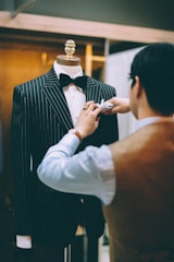 A tailor is adjusting a pinstripe suit on a mannequin. The suit is black with white pinstripes and is complemented by a black bow tie. The tailor is wearing a watch on their left wrist and is focused on perfecting the details of the suit. The background is softly blurred, giving attention to the tailor's meticulous work.