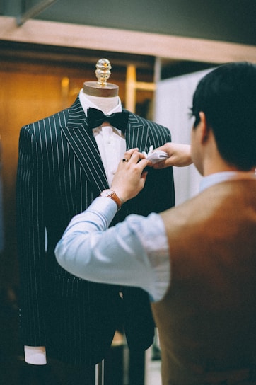 A tailor is adjusting a pinstripe suit on a mannequin. The suit is black with white pinstripes and is complemented by a black bow tie. The tailor is wearing a watch on their left wrist and is focused on perfecting the details of the suit. The background is softly blurred, giving attention to the tailor's meticulous work.