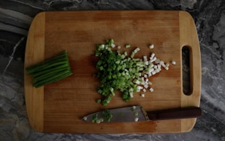 A wooden cutting board with chopped green onions scattered on top. To the side, there is a pile of unchopped green onions and a knife with a dark handle resting on the board. The surface beneath the board has a dark, marbled pattern.