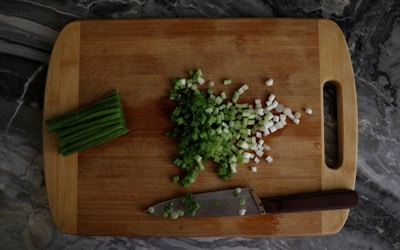 A wooden cutting board with chopped green onions scattered on top. To the side, there is a pile of unchopped green onions and a knife with a dark handle resting on the board. The surface beneath the board has a dark, marbled pattern.