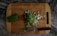 A wooden cutting board with chopped green onions scattered on top. To the side, there is a pile of unchopped green onions and a knife with a dark handle resting on the board. The surface beneath the board has a dark, marbled pattern.