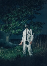 A person wearing a gas mask and protective suit stands in a grassy area at night. The figure holds a device emitting smoke, surrounded by tall grasses and a tree.