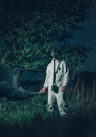 A person wearing a gas mask and protective suit stands in a grassy area at night. The figure holds a device emitting smoke, surrounded by tall grasses and a tree.