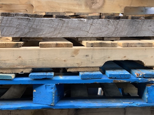 A stack of wooden pallets with different textures and colors. The top pallet appears weathered with a mix of gray and tan shades, while the lower pallet is painted blue and shows signs of wear and tear.