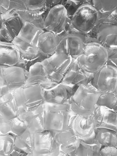 A collection of clear ice cubes arranged in a close-up view, showing the smooth and somewhat irregular shapes of the cubes. The ice has a glossy finish with visible reflections and occasional air bubbles trapped within.