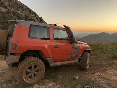 A rugged orange SUV with large tires is parked on a dirt path near rocky terrain. The vehicle features a spare tire on the back and a 4x4 emblem on the side. The image captures a scenic background with grassy hills, distant mountains, and a sunset sky with soft pastel hues.