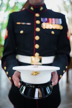 A person is dressed in a formal military uniform adorned with medals and badges. The individual holds a white peaked cap with a black visor. The uniform is buttoned and features a high collar, with gold buttons and colorful ribbons on the chest.