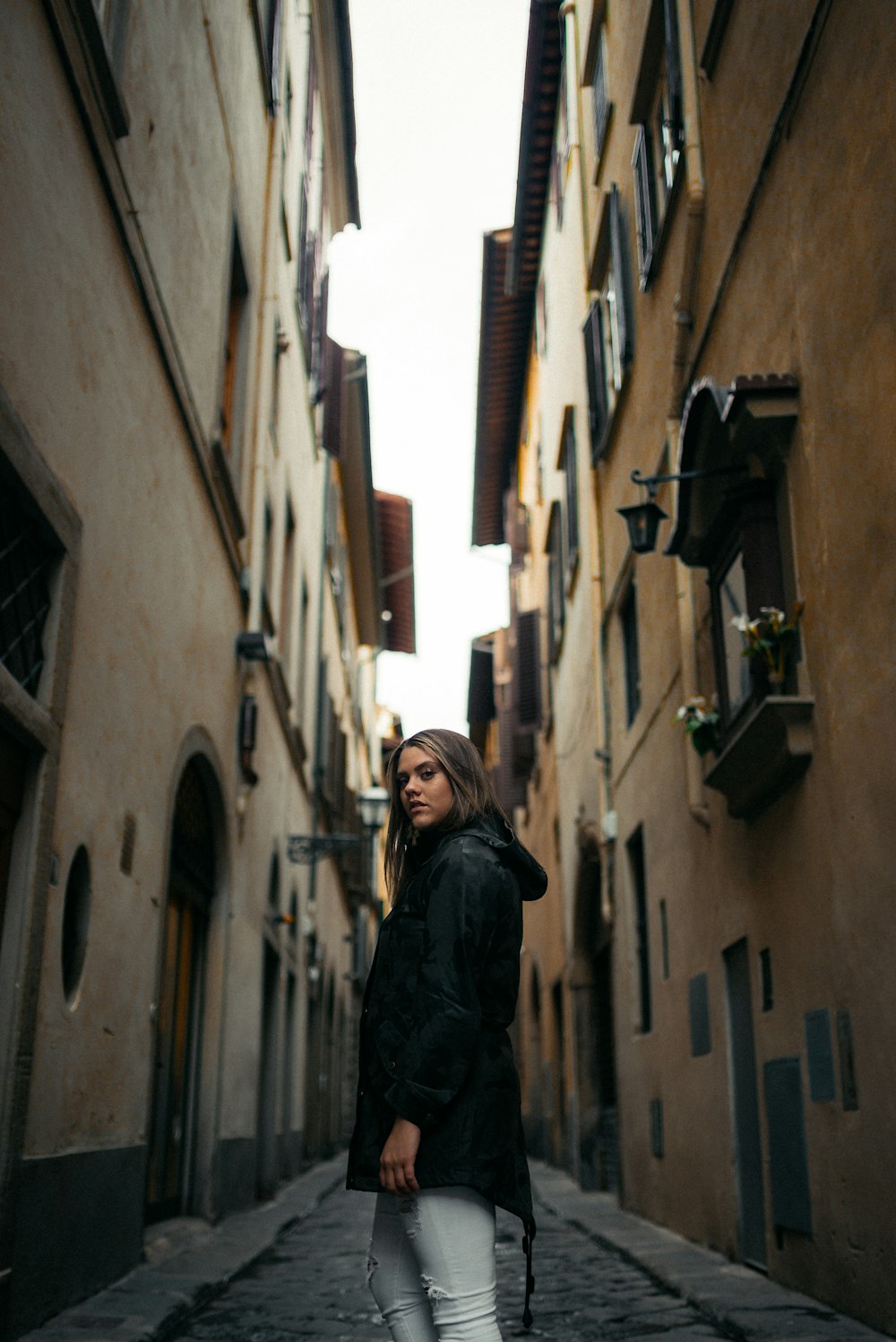Femme en veste noire debout entre les bâtiments pendant la journée