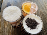 Three glasses filled with different beverages are arranged on a wooden table. One glass contains a frothy white drink, another has a citrus-based drink with a slice of orange and ice cubes, and the last glass holds a creamy beverage topped with crushed dark brown bits.