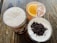Three glasses filled with different beverages are arranged on a wooden table. One glass contains a frothy white drink, another has a citrus-based drink with a slice of orange and ice cubes, and the last glass holds a creamy beverage topped with crushed dark brown bits.