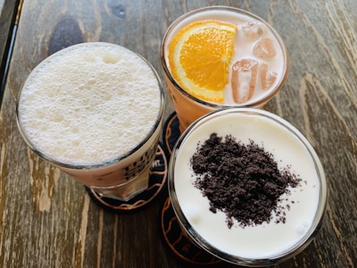 Three glasses filled with different beverages are arranged on a wooden table. One glass contains a frothy white drink, another has a citrus-based drink with a slice of orange and ice cubes, and the last glass holds a creamy beverage topped with crushed dark brown bits.