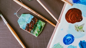 Several paintbrushes are arranged on a textured surface alongside a rectangular palette filled with various blobs of paint. The palette features colors like brown, blue, and green. A small, vibrant painting on paper shows a landscape with mountains, sky, and greenery, connected by a string as if part of a tag.