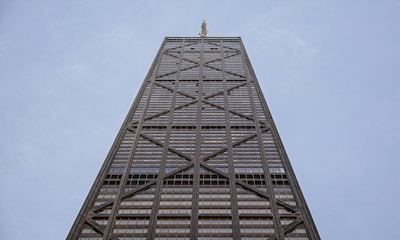 A tall skyscraper with a steel and glass facade, featuring a series of diagonal cross braces. The building reaches upward into a clear blue sky, showcasing modern architectural design and engineering.