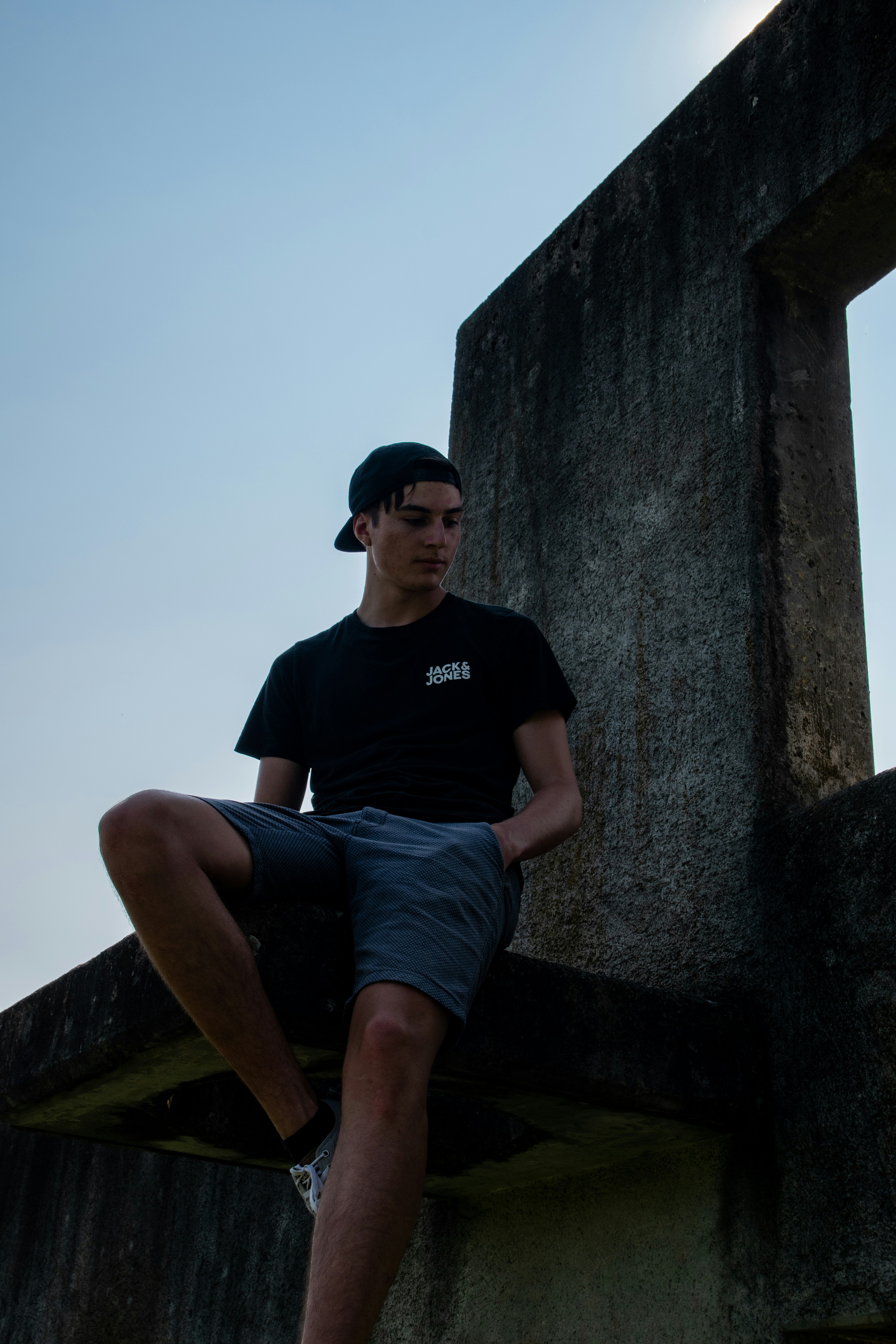 man in black crew neck t-shirt and blue denim shorts sitting on concrete wall during