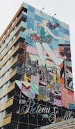 A building features a large, colorful mural depicting a woman in a ballet pose, seemingly leaping into the air with the city skyline in the background. The mural consists of vivid blocks of color and various artistic patterns.