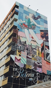 A building features a large, colorful mural depicting a woman in a ballet pose, seemingly leaping into the air with the city skyline in the background. The mural consists of vivid blocks of color and various artistic patterns.