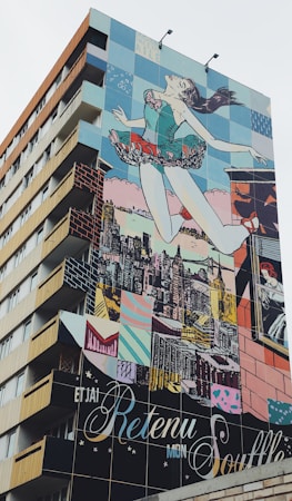 A building features a large, colorful mural depicting a woman in a ballet pose, seemingly leaping into the air with the city skyline in the background. The mural consists of vivid blocks of color and various artistic patterns.