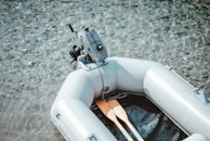 A small inflatable boat with a motor is floating on clear, shallow water with visible pebbles underneath. Two paddles are placed inside the boat, ready for use.