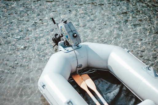 A small inflatable boat with a motor is floating on clear, shallow water with visible pebbles underneath. Two paddles are placed inside the boat, ready for use.