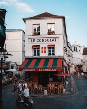 A charming, old-fashioned restaurant named 'Le Consulat' is located at a street corner with a distinctive red, green, and white awning. The establishment features outdoor seating with empty wooden chairs and tables arranged on the cobblestone pavement. Several people are present, including a motorcyclist wearing a helmet and two individuals seated at a table. The setting is a quaint, picturesque street with a clear blue sky overhead and classic European architecture in the background.