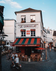 A charming, old-fashioned restaurant named 'Le Consulat' is located at a street corner with a distinctive red, green, and white awning. The establishment features outdoor seating with empty wooden chairs and tables arranged on the cobblestone pavement. Several people are present, including a motorcyclist wearing a helmet and two individuals seated at a table. The setting is a quaint, picturesque street with a clear blue sky overhead and classic European architecture in the background.