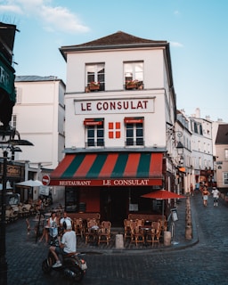 A charming, old-fashioned restaurant named 'Le Consulat' is located at a street corner with a distinctive red, green, and white awning. The establishment features outdoor seating with empty wooden chairs and tables arranged on the cobblestone pavement. Several people are present, including a motorcyclist wearing a helmet and two individuals seated at a table. The setting is a quaint, picturesque street with a clear blue sky overhead and classic European architecture in the background.