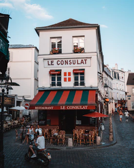 A charming, old-fashioned restaurant named 'Le Consulat' is located at a street corner with a distinctive red, green, and white awning. The establishment features outdoor seating with empty wooden chairs and tables arranged on the cobblestone pavement. Several people are present, including a motorcyclist wearing a helmet and two individuals seated at a table. The setting is a quaint, picturesque street with a clear blue sky overhead and classic European architecture in the background.