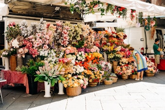 A colorful flower market with a variety of blooms displayed in baskets and pots on the pavement. Two people are interacting with the flowers; one is examining a bouquet while another stands near a white van in the background. The stall is covered with a canopy and surrounded by vibrant assortments of flowers including roses, lilies, and daisies, creating a lively and bustling atmosphere.