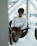 A person with glasses is sitting comfortably in a modern chair by a large window, reading a book. They are wearing a white long-sleeve shirt with the words 'Redefine Health' printed on it and gray pants. Their legs are extended and resting on a small table, with shoes facing outward. The setting appears to be indoors with natural light coming through the window.