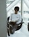 A person with glasses is sitting comfortably in a modern chair by a large window, reading a book. They are wearing a white long-sleeve shirt with the words 'Redefine Health' printed on it and gray pants. Their legs are extended and resting on a small table, with shoes facing outward. The setting appears to be indoors with natural light coming through the window.