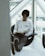 A person with glasses is sitting comfortably in a modern chair by a large window, reading a book. They are wearing a white long-sleeve shirt with the words 'Redefine Health' printed on it and gray pants. Their legs are extended and resting on a small table, with shoes facing outward. The setting appears to be indoors with natural light coming through the window.