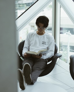A person with glasses is sitting comfortably in a modern chair by a large window, reading a book. They are wearing a white long-sleeve shirt with the words 'Redefine Health' printed on it and gray pants. Their legs are extended and resting on a small table, with shoes facing outward. The setting appears to be indoors with natural light coming through the window.
