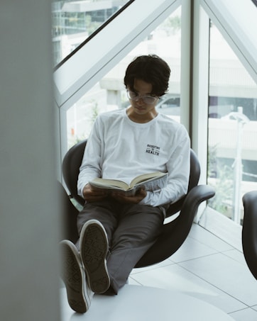 A person with glasses is sitting comfortably in a modern chair by a large window, reading a book. They are wearing a white long-sleeve shirt with the words 'Redefine Health' printed on it and gray pants. Their legs are extended and resting on a small table, with shoes facing outward. The setting appears to be indoors with natural light coming through the window.
