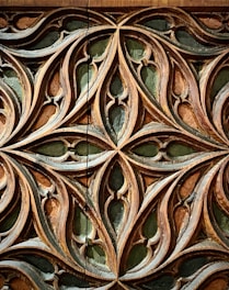 Intricately carved wooden pattern featuring interlocking curves and pointed shapes. The design showcases an elaborate lattice with symmetrical motifs, highlighted by a contrast between the wood's natural tones and a dark green background.