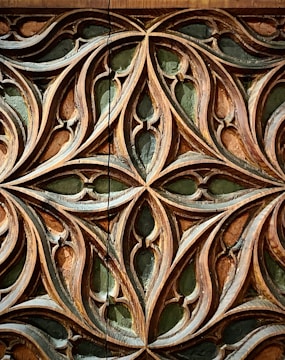 Intricately carved wooden pattern featuring interlocking curves and pointed shapes. The design showcases an elaborate lattice with symmetrical motifs, highlighted by a contrast between the wood's natural tones and a dark green background.