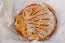 A round, rustic sandwich with a decorative imprint on the top, resting on crumpled white paper. The sandwich appears to be filled with slices of cured meat.