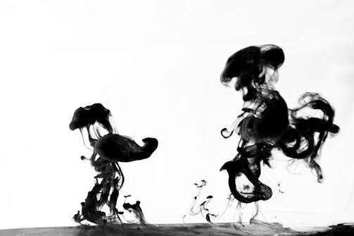 Black ink swirls and spreads in clear liquid, creating abstract shapes against a bright background. The fluid forms intricate, flowing patterns with a mix of thick and thin tendrils as the ink disperses.
