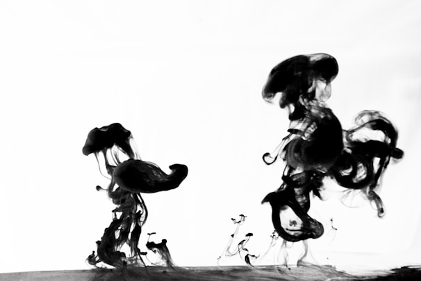 Black ink swirls and spreads in clear liquid, creating abstract shapes against a bright background. The fluid forms intricate, flowing patterns with a mix of thick and thin tendrils as the ink disperses.