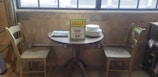 A small wooden dining table set against a rustic concrete wall with an industrial vibe. There are two wooden chairs placed on either side of the table, which has a stack of plates and a menu card indicating a discounted price for the set. The setting is by a large window, allowing natural light to illuminate the scene.