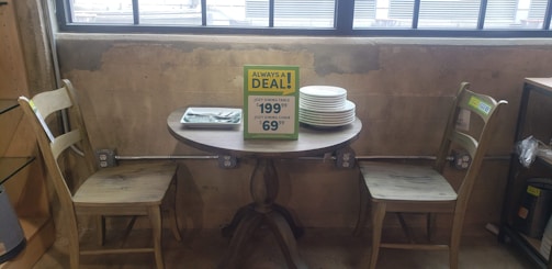A small wooden dining table set against a rustic concrete wall with an industrial vibe. There are two wooden chairs placed on either side of the table, which has a stack of plates and a menu card indicating a discounted price for the set. The setting is by a large window, allowing natural light to illuminate the scene.