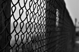 A close-up view of a metal chain-link fence supported by wooden posts. The image is captured in black and white, giving it a moody and somber feel. The background is blurred and out of focus, emphasizing the fence's texture and pattern.