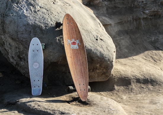 Two longboards are resting against a large, textured rock on a rugged, sandy surface. One board has a natural wood finish with an emblem, while the other has a lighter, textured grip.