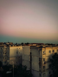 A group of multi-story residential buildings with a flat, muted sunset sky above. The architecture is utilitarian with a slightly worn appearance. Some windows are visible, and there is sparse vegetation around the buildings.