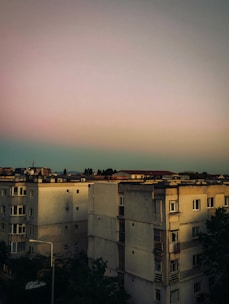 A group of multi-story residential buildings with a flat, muted sunset sky above. The architecture is utilitarian with a slightly worn appearance. Some windows are visible, and there is sparse vegetation around the buildings.