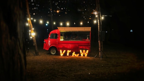 A vintage red food truck is parked in a dimly lit outdoor setting. Large illuminated letters spelling 'vr&w' are placed in front of the truck. String lights are hanging above, casting a warm glow. The environment appears to be a wooded area at night, contributing to a cozy atmosphere.