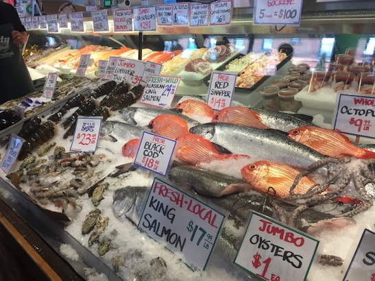 A well-stocked seafood market display featuring a variety of fresh fish and shellfish on ice. Visible species include Alaskan King Salmon, Red Snapper, Black Cod, lobsters, squid, oysters, and octopus. Handwritten signs display prices and product labels. The display is well-organized, with seafood items neatly arranged in rows.